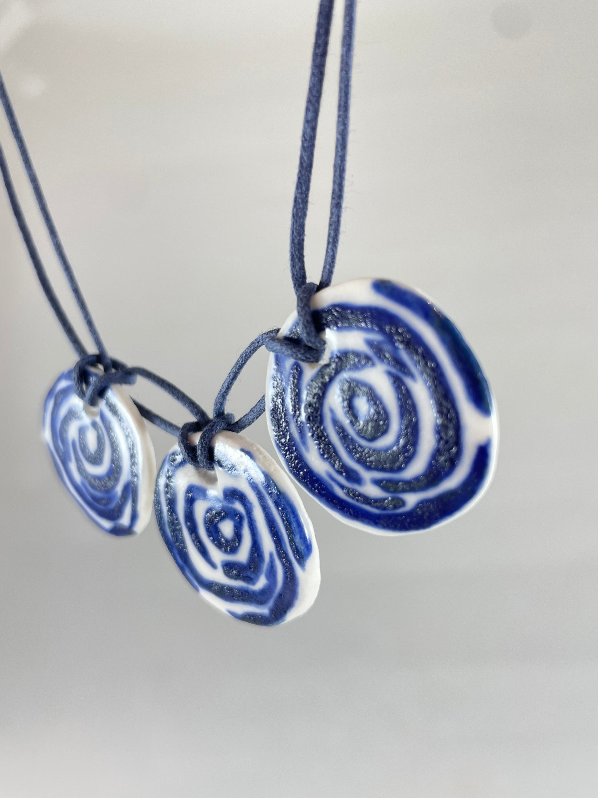 Necklace with three circular porcelain pendants on navy blue coloured thread, hanging in front of a white background. Blue rose pattern painted on white porcelain.  Photographed from right side to show thin profile of porcelain discs