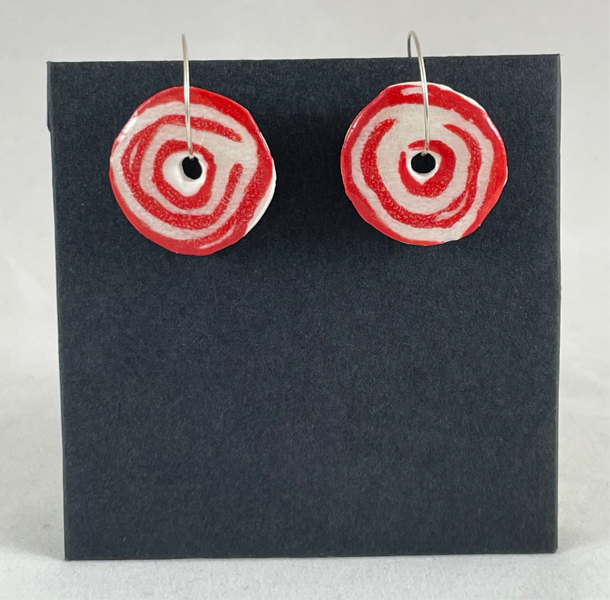 A pair of red and white porcelain earrings hanging on black card. Simple rose pattern with loosely painted red circles on white porcelain.