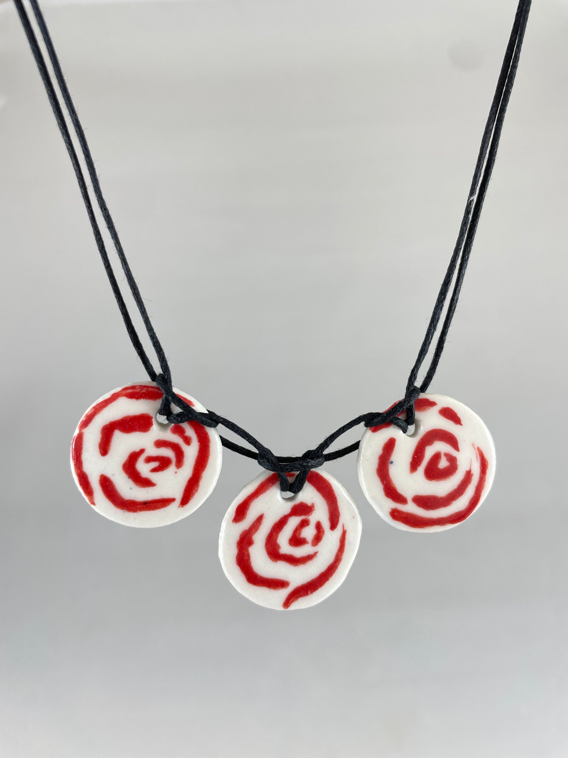 Necklace with three circular porcelain pendants on black coloured thread, hanging in front of a white background. Red rose pattern painted on white porcelain.  