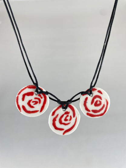 Necklace with three circular porcelain pendants on black coloured thread, hanging in front of a white background. Red rose pattern painted on white porcelain.  