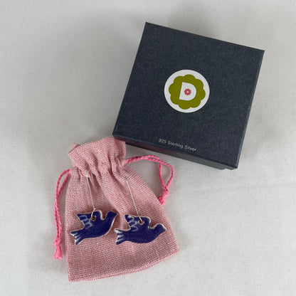 A pair of lavender porcelain earrings in the shape of flying dove. Photographed on a linen pouch next to jewellery box.
