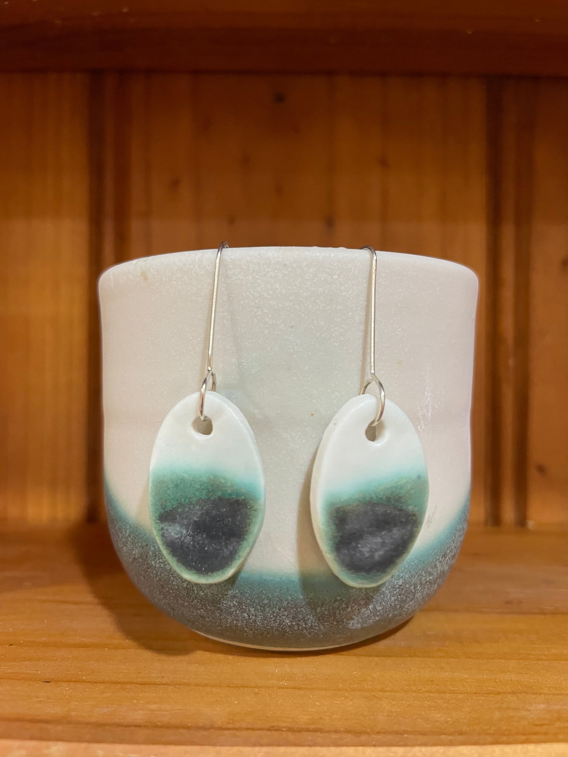 Pair of emerald green and white oval shaped porcelain earrings. Top half white and bottom half textured green. Sterling silver hooks. Pictured in front of a white and green ceramic cup 