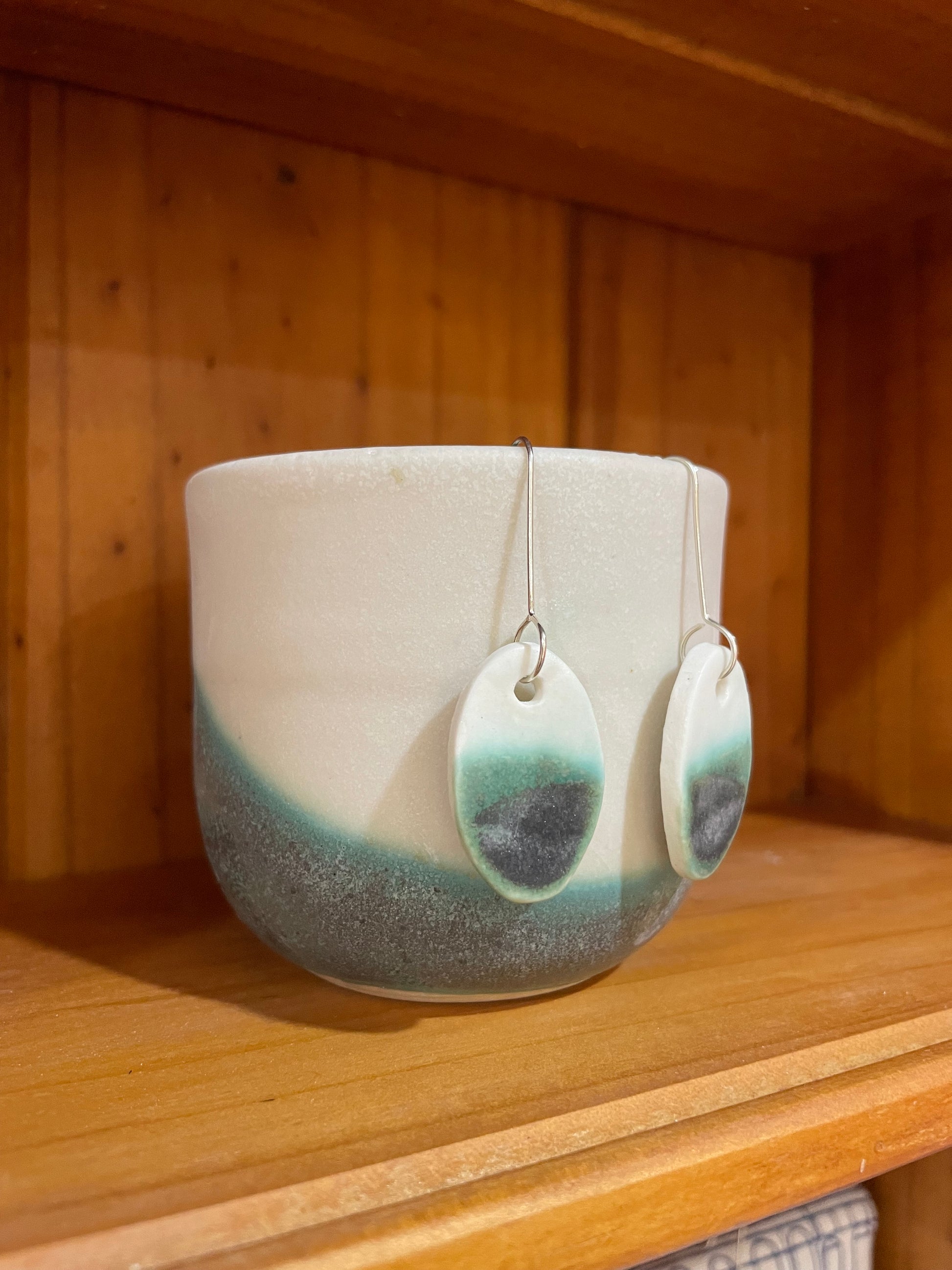 Pair of emerald green and white oval shaped porcelain earrings. Top half white and bottom half textured green. Sterling silver hooks. Pictured in front of a white and green ceramic cup, side view