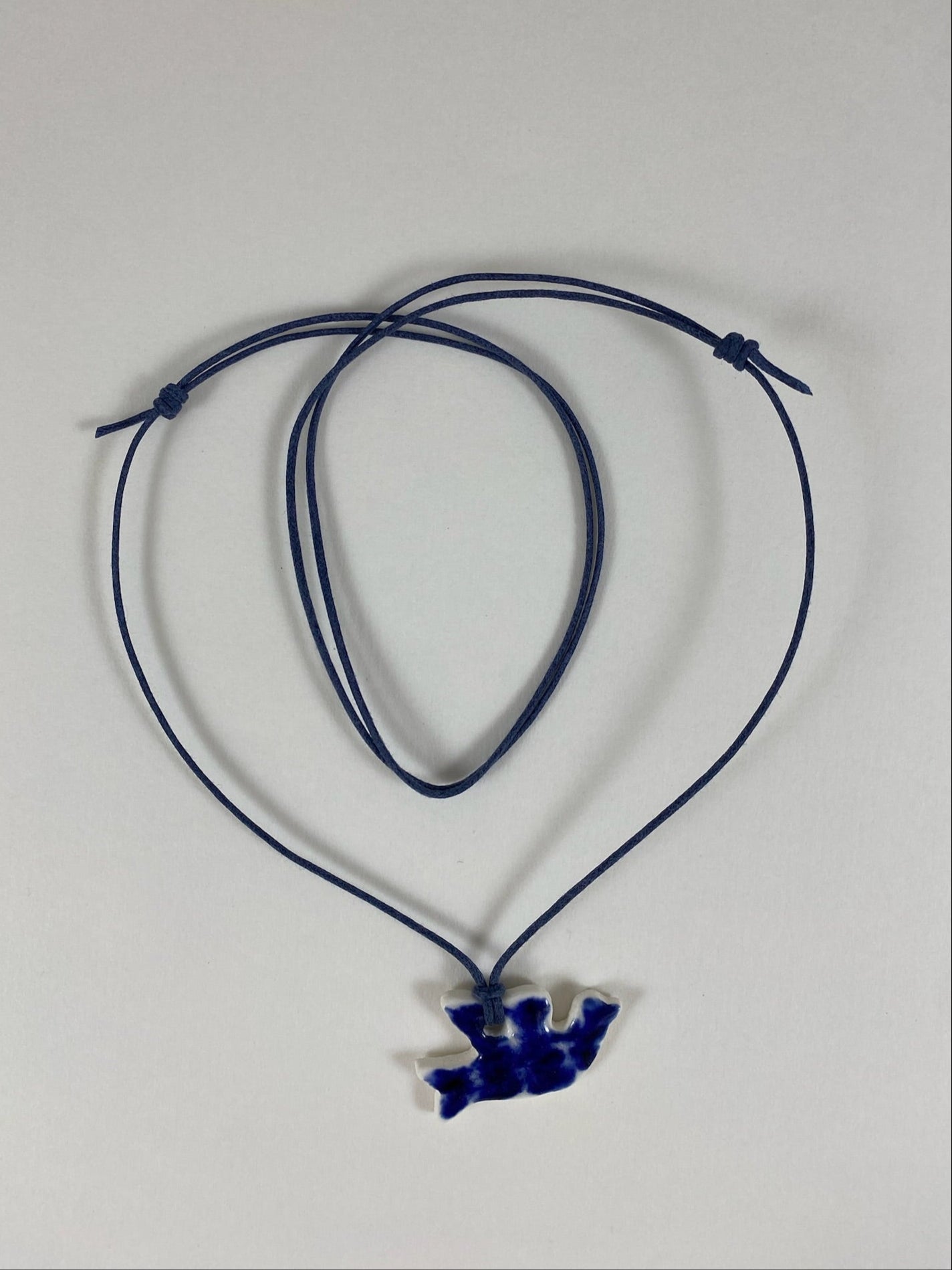 Necklace laid flat on white background. Fine black thread with dove shaped pendant in dark blue. Edges of pendant are white. Pendant is attached with simple larks head knot.