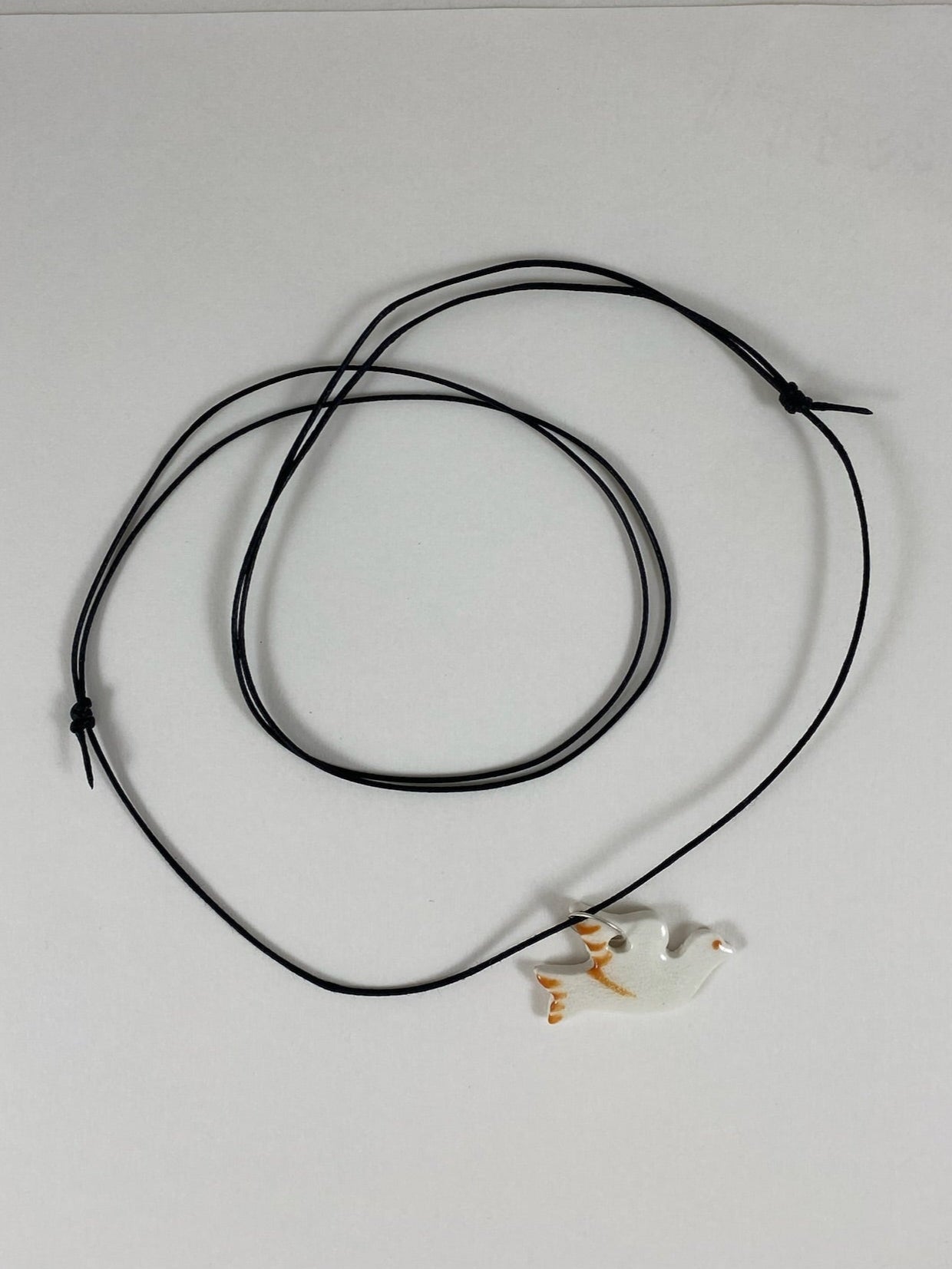 Necklace laid flat on white background. Fine black thread with dove shaped pendant in white with orange wing and tail. Pendant is attached with small sterling silver jump ring.