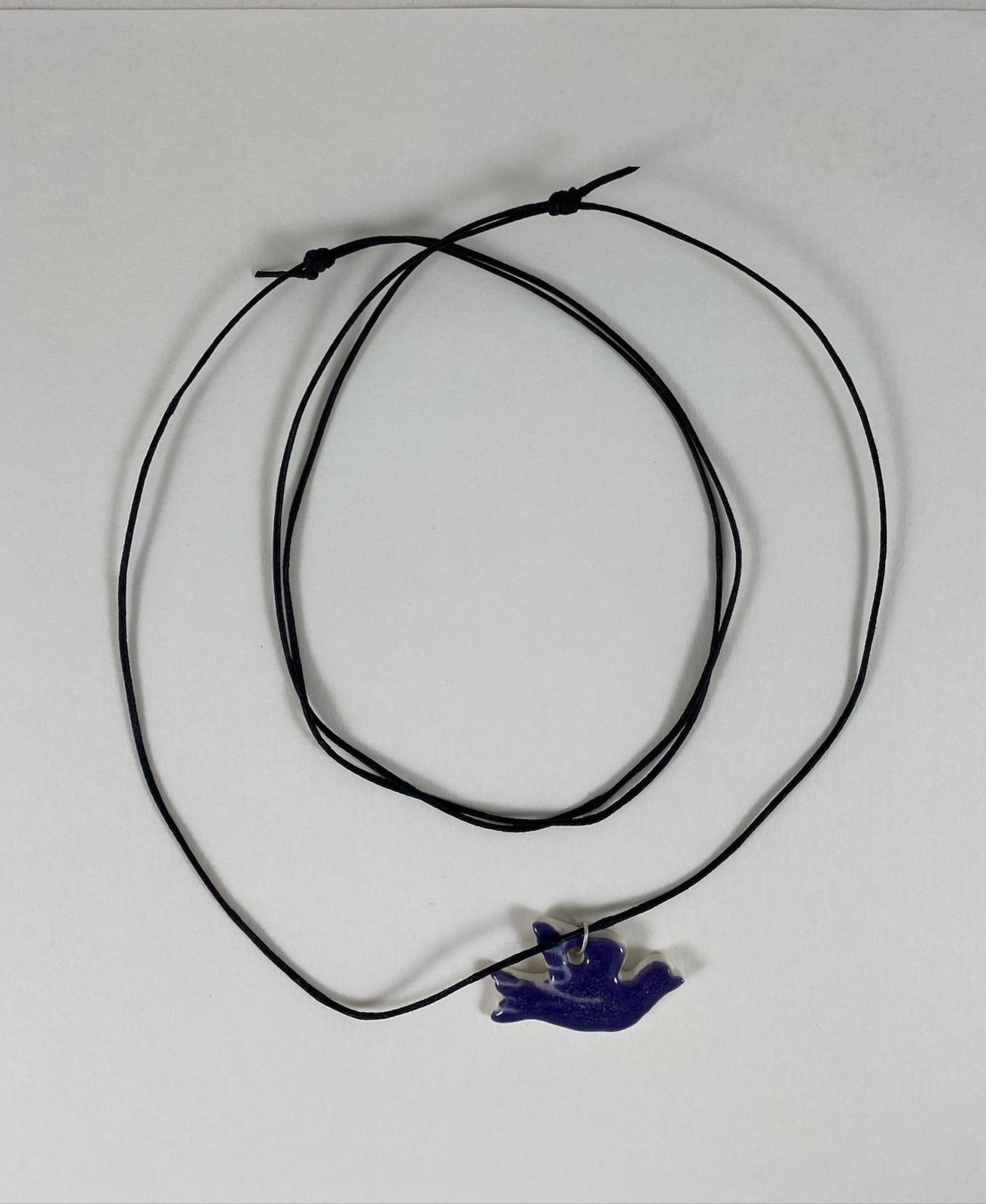 Necklace laid flat on white background. Fine black thread with dove shaped pendant in dark lavender purple. Fine white etchings on wing and tail. Pendant is attached with sterling silver jump ring.