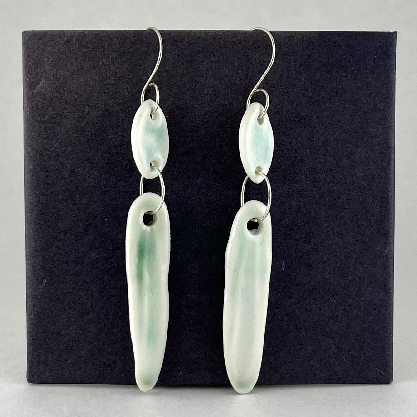 Pair of pale green porcelain earrings on a black card. Short curved silver earring hooks. Each earring has a small leaf shaped pendant, connected to a long thin leaf shaped pendant with a shiny round silver jump ring.