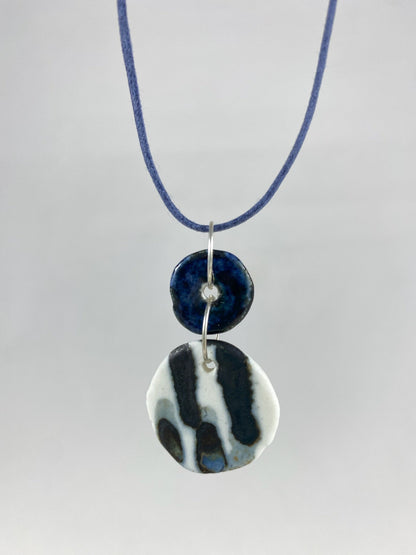 Necklace hanging against white background. Fine navy blue string with two circular porcelain pendants attached with silver jump rings. One pendant is small and navy blue and the larger pendant is white with navy glaze drips that looks like watercolour paint.