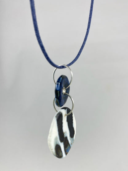 Necklace hanging against white background. Fine navy blue string with two circular porcelain pendants attached with silver jump rings. One pendant is small and navy blue and the larger pendant is white with navy glaze drips that looks like watercolour paint. Side view. Pendants are thin porcelain.