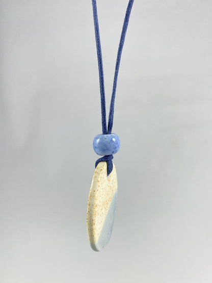 Necklace hanging against white background. Fine navy blue thread with pendant in cream with orange flecks and pale blue. A small pale blue porcelain bead sits above the pendant. Side view. The porcelain pendant is thin.