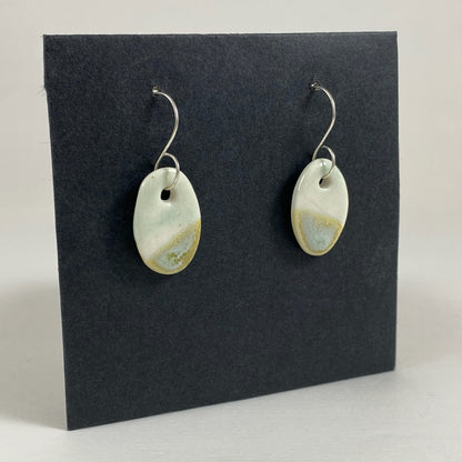 Fine rounded silver hooks with oval shaped pendants in white porcelain. Bottom half dipped asymmetrically in glaze speckled with light green, light brown and pale blue.  Photographed on a black background from the left hand side. The porcelain is thin, approximately 2 mm thick. 