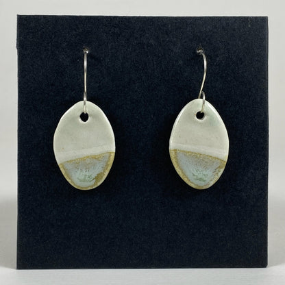 Fine rounded silver hooks with oval shaped pendants in white porcelain. Bottom half dipped asymmetrically in glaze speckled with light green, light brown and pale blue.  Photographed on a black background.