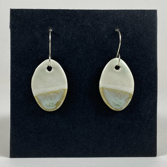 Fine rounded silver hooks with oval shaped pendants in white porcelain. Bottom half dipped asymmetrically in glaze speckled with light green, light brown and pale blue.  Photographed on a black background.