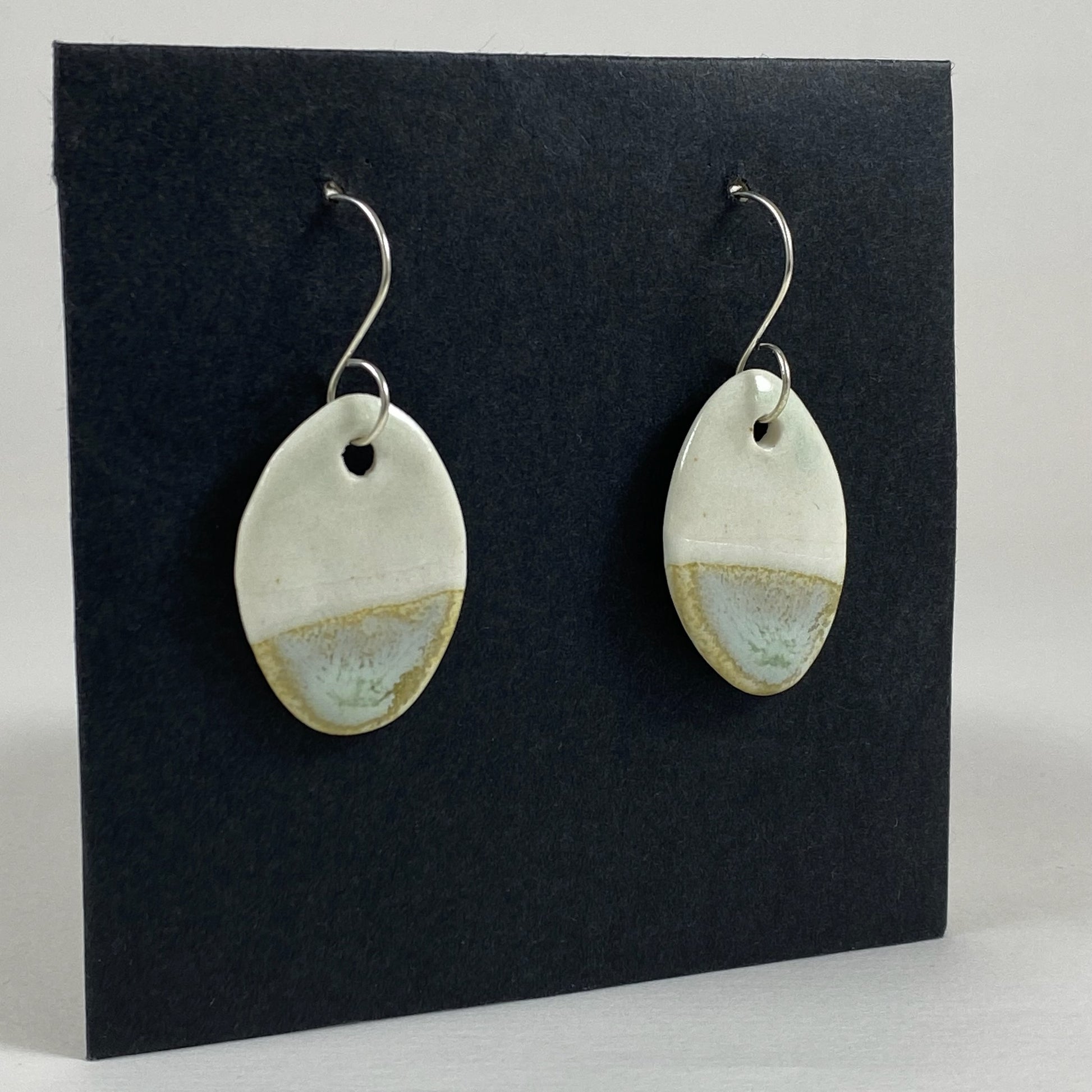 Fine rounded silver hooks with oval shaped pendants in white porcelain. Bottom half dipped in glaze speckled with light green, light brown and pale blue.  Photographed on a black background from the left hand side. The porcelain is thin, approximately 3 to 4 mm thick. 