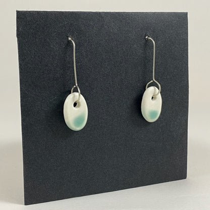Fine silver hooks with oval shaped pendants in white porcelain. Bottom half  dipped in mint green shiny glaze, which fades  to white at the edges for a soft look.  Photographed on a black background from the left hand side. The porcelain is thin, approximately 2 mm thick. 