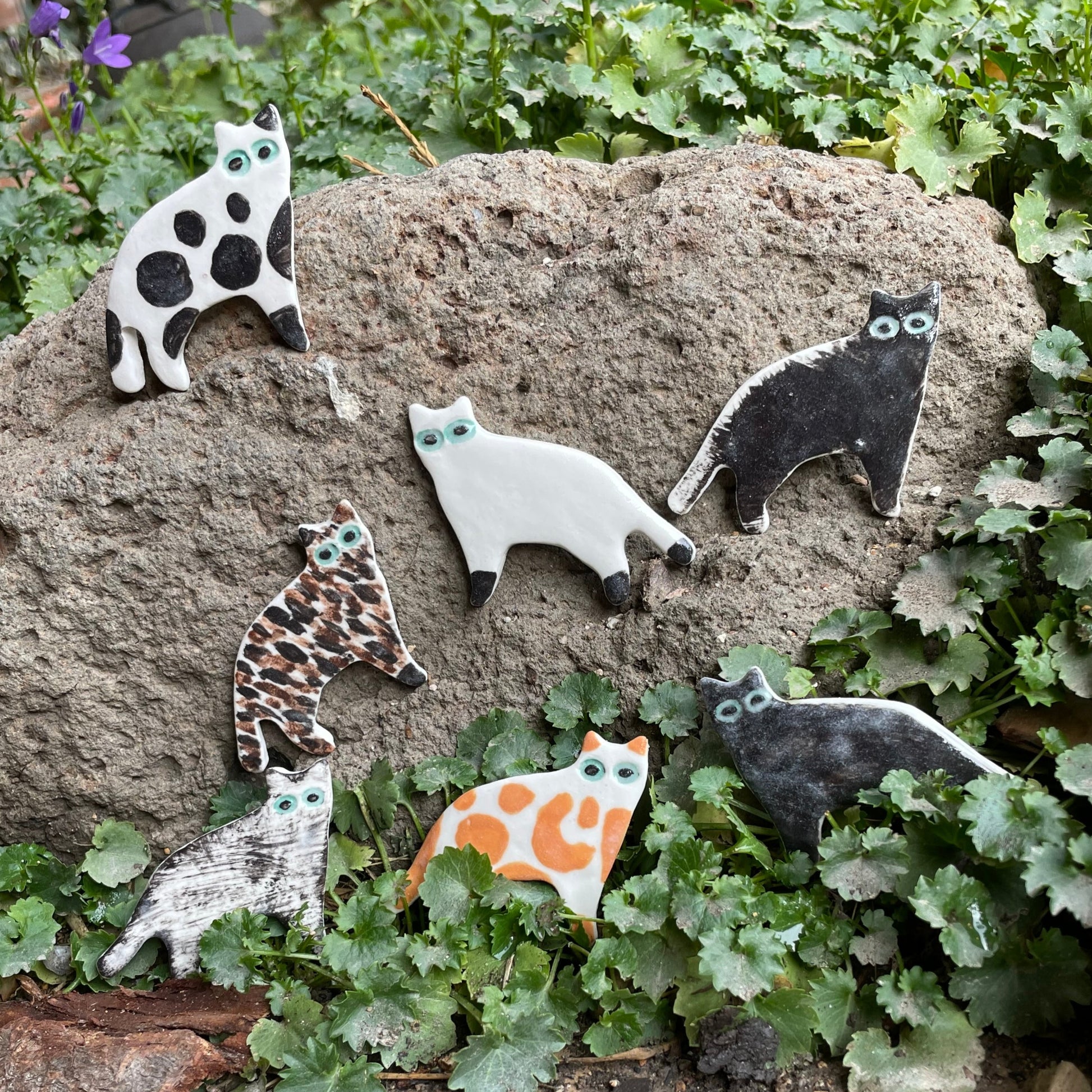 Seven porcelain cats in a range of colours and patterns. Photographed outside in garden. Arranged leaning against a large volcanic rock with foliage around.