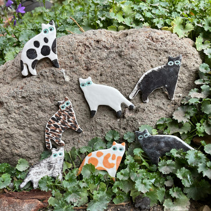 Seven porcelain cats in a range of colours and patterns. Photographed outside in garden. Arranged leaning against a large volcanic rock with foliage around.