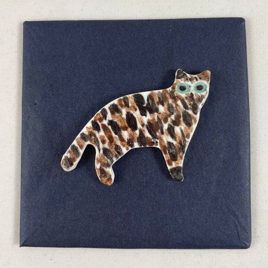 Cat brooch in standing position. Brown and black brush strokes on white porcelain. Effect is like a tiger striped cat. Large green eyes. Photographed on black background.
