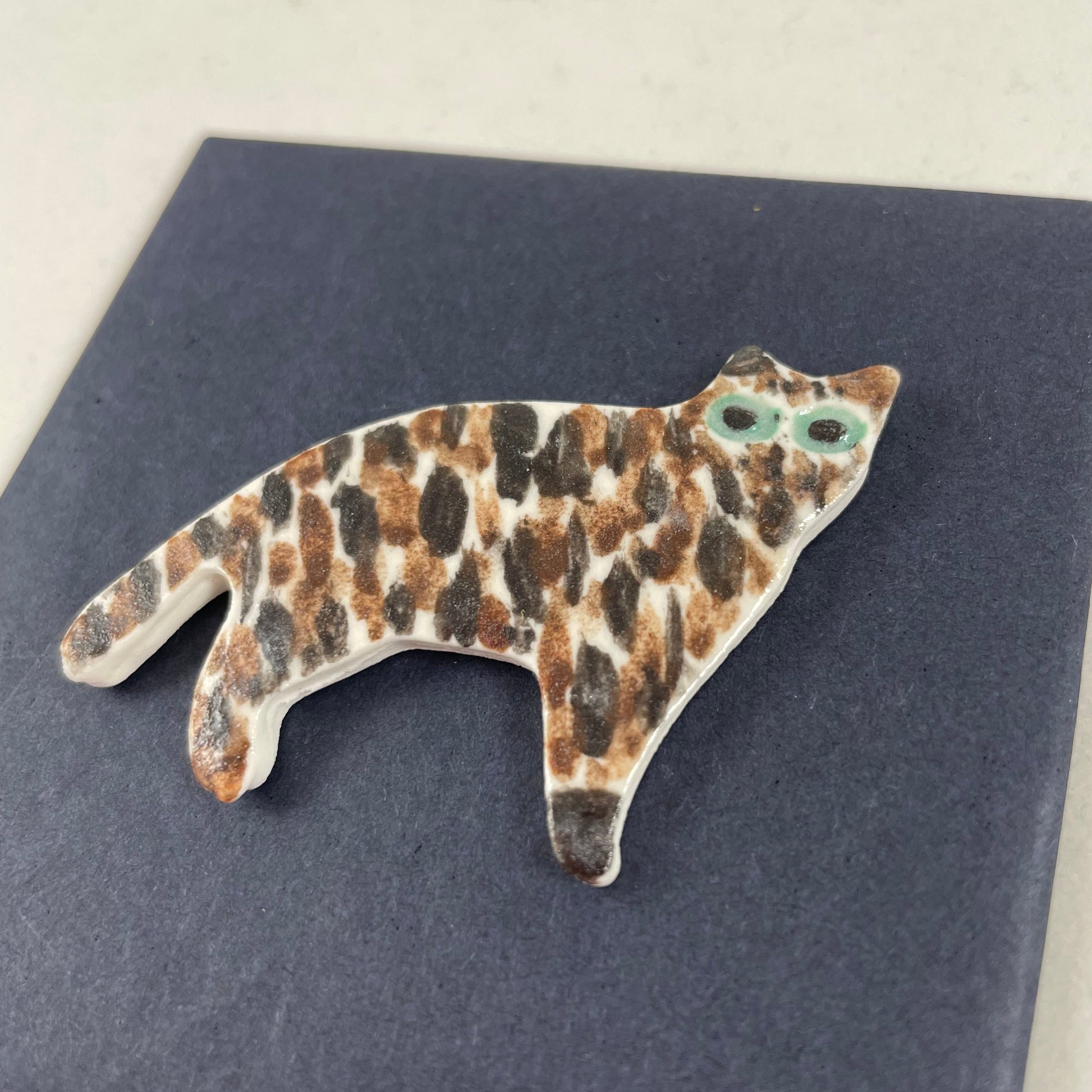 Cat brooch in standing position. Brown and black brush strokes on white porcelain. Effect is like a tiger striped cat. Large green eyes. Photographed on black background from right hand side. Made of thin porcelain.