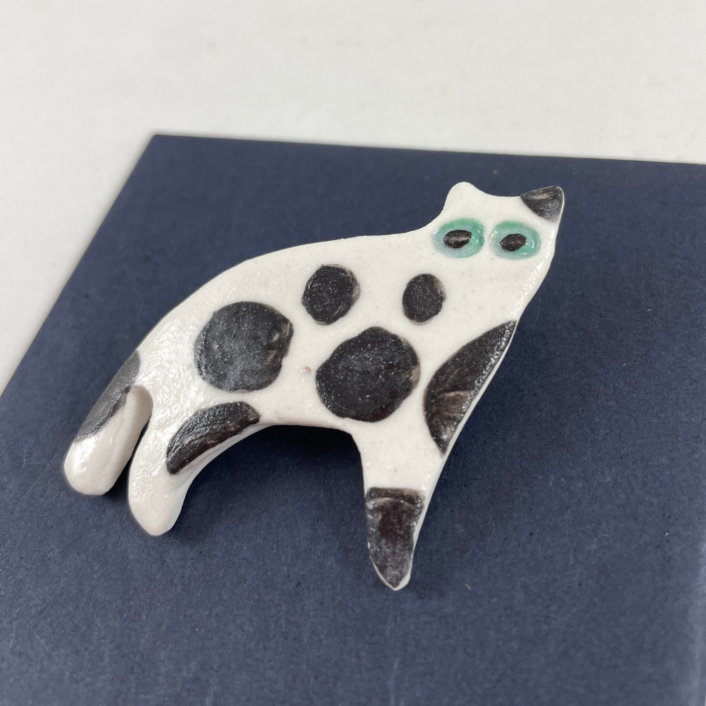 White porcelain cat with large irregular black spots and patches. Large green eyes. Photographed flat on a black background from right hand side. Made of thin porcelain