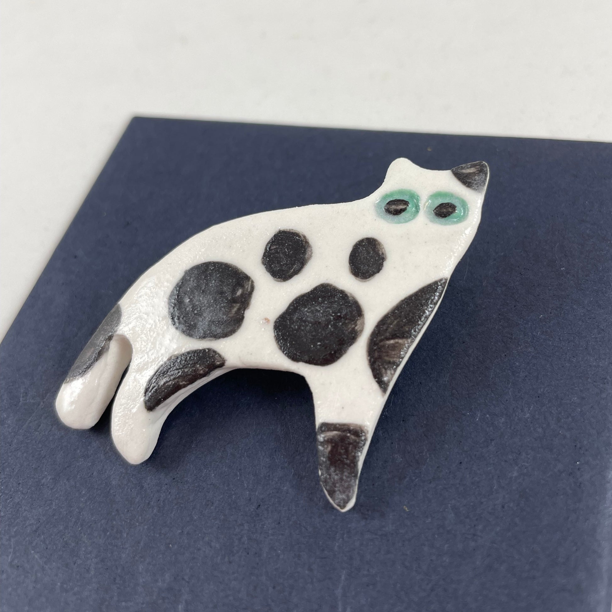White porcelain cat with large irregular black spots and patches. Large green eyes. Photographed flat on a black background from right hand side. Made of thin porcelain