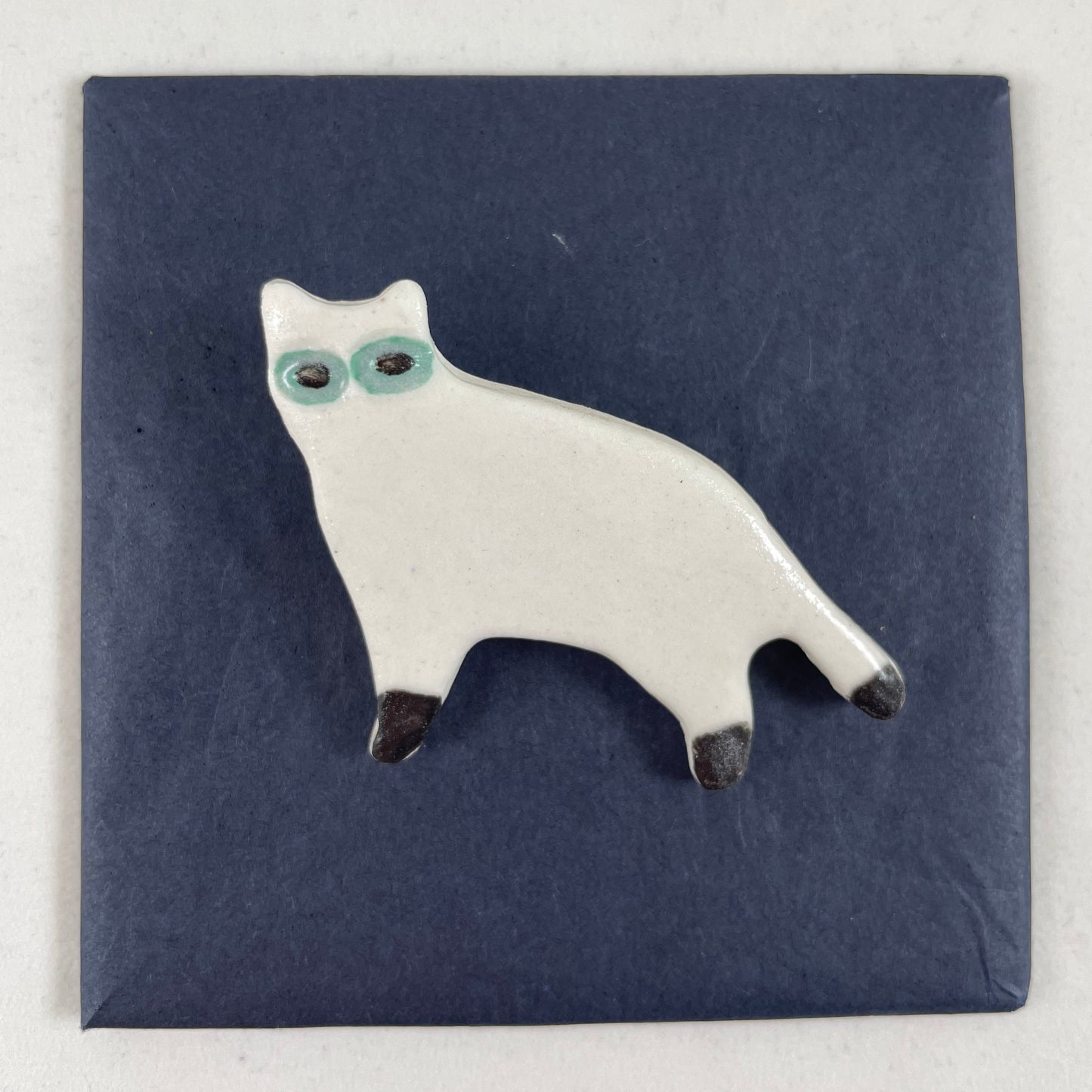 White porcelain cat standing with black feet and tip of tail. Large green eyes. Photographed flat on a black background.