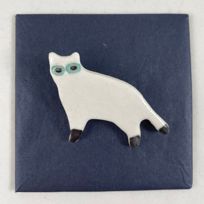 White porcelain cat standing with black feet and tip of tail. Large green eyes. Photographed flat on a black background.