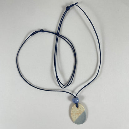 Necklace laid flat on white background. Fine navy blue string with one porcelain pendant and above it a small light blue porcelain bead. The pendant is oval and half cream with tiny orange flecks, and half light blue/lilac. 