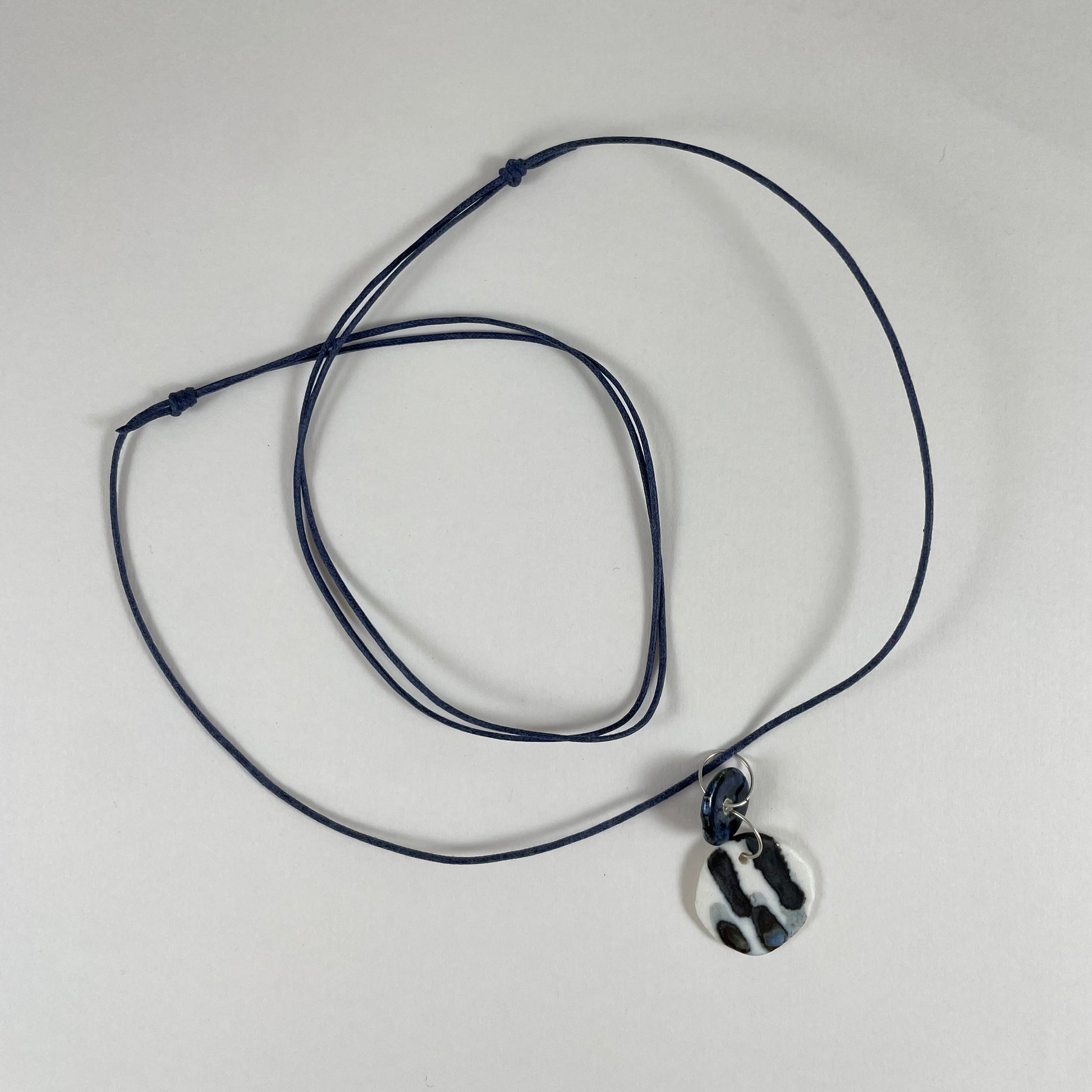 Necklace laid flat on white background. Fine black string with two circular porcelain pendants attached with silver jump rings. One pendant is small and navy blue and the larger pendant is white with navy glaze drips that looks like watercolour paint.