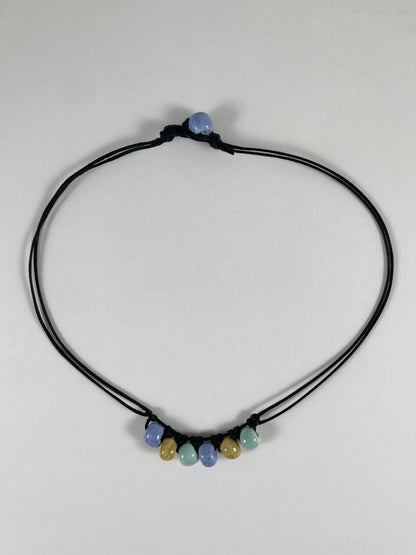 Short necklace laid flat on white background. Two strands of fine black string with six porcelain beads attached using simple macrame knots. Two beads are light blue, two are gold and two are light mint green. They are arranged in an alternating order. One end of necklace has a blue bead attached and the other end has a small loop for the bead to attach to.