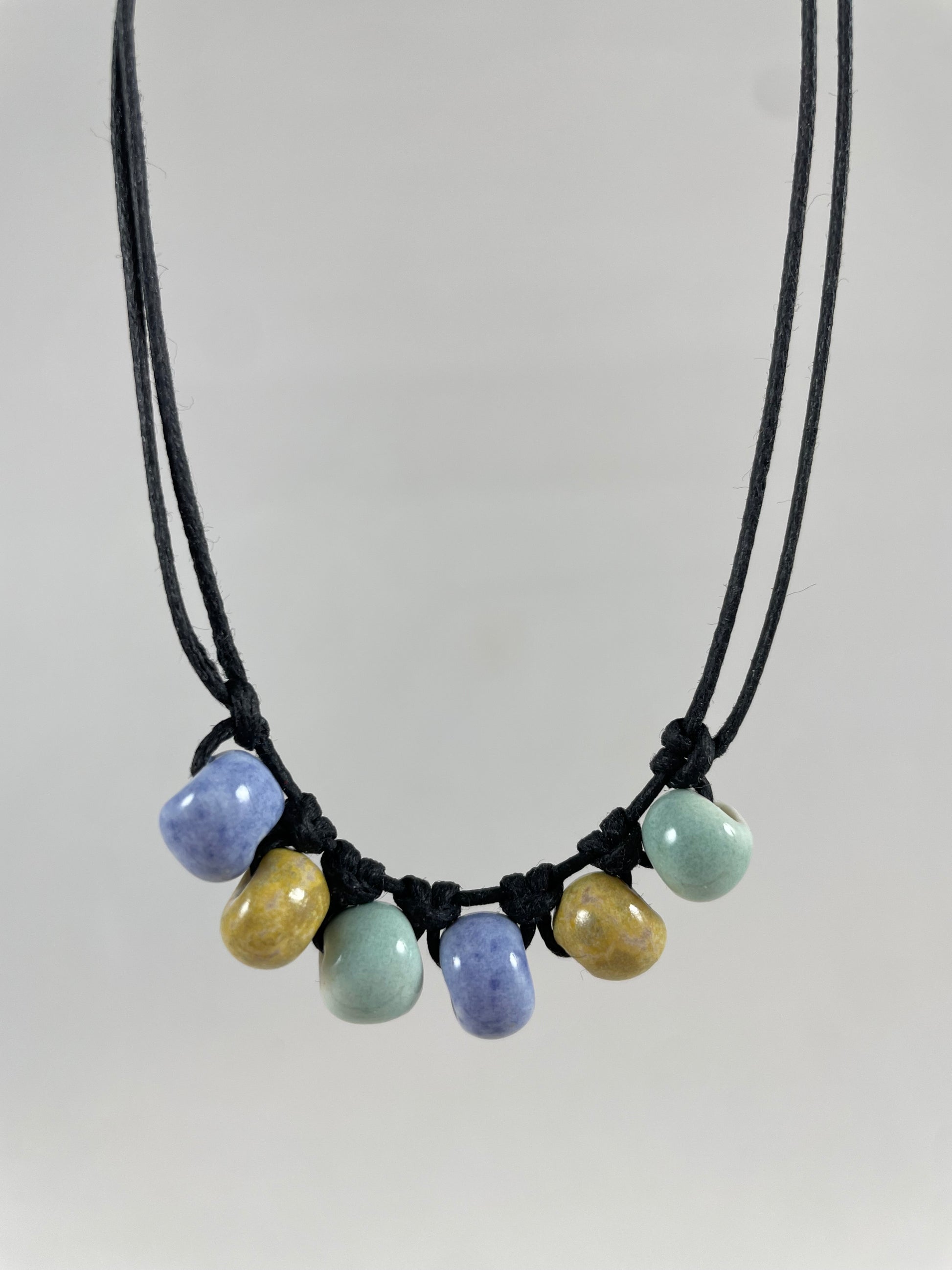Short necklace hanging against white background. Two strands of fine black string with six porcelain beads attached using simple macrame knots. Two beads are light blue, two are gold and two are light mint green. They are arranged in an alternating order.