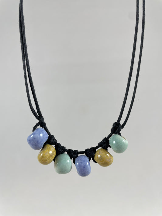 Short necklace hanging against white background. Two strands of fine black string with six porcelain beads attached using simple macrame knots. Two beads are light blue, two are gold and two are light mint green. They are arranged in an alternating order.