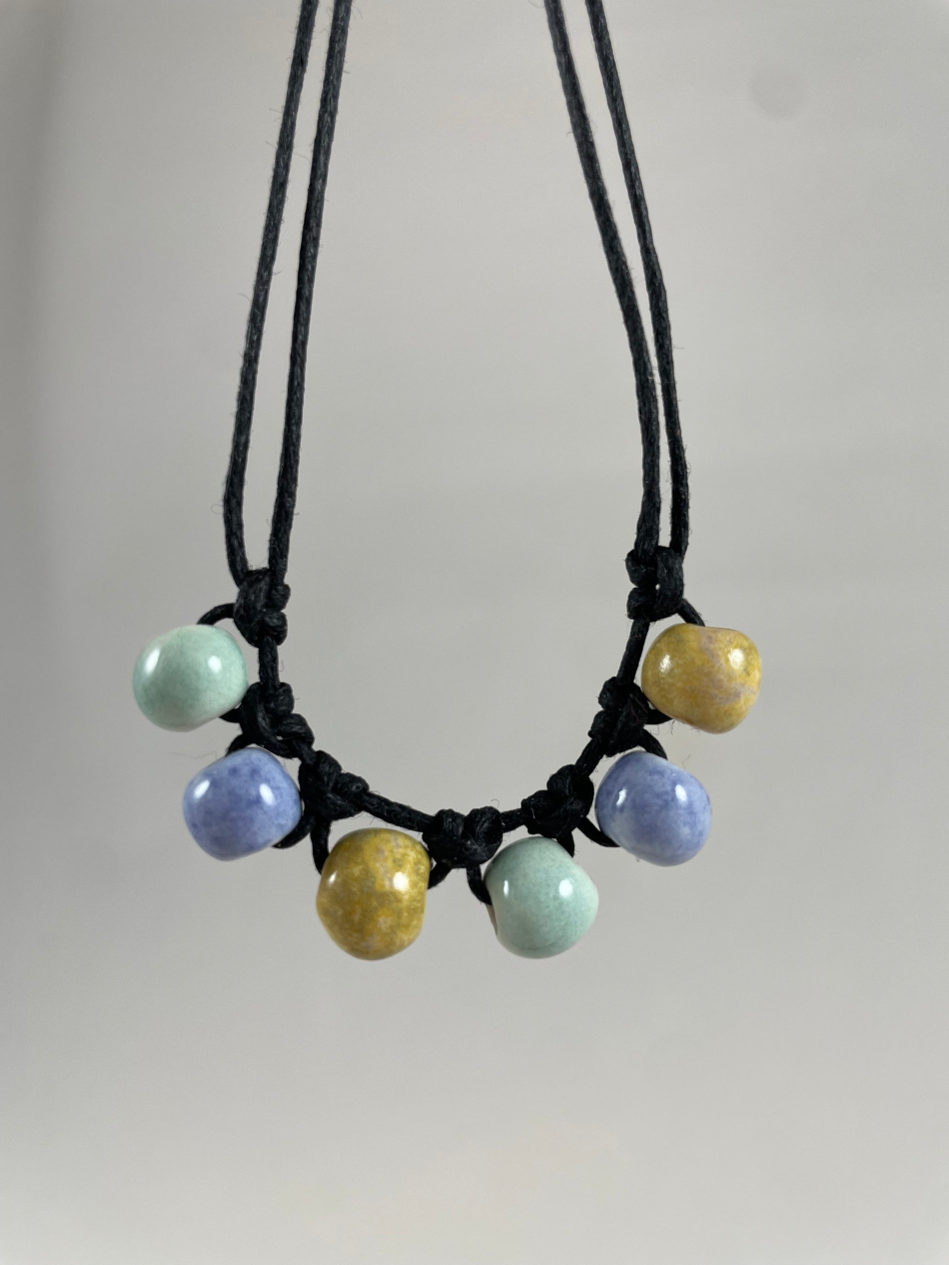 Short necklace hanging against white background. Two strands of fine black string with six porcelain beads attached using simple macrame knots. Two beads are light blue, two are gold and two are light mint green. They are arranged in an alternating order.