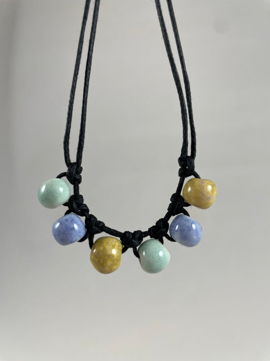 Short necklace hanging against white background. Two strands of fine black string with six porcelain beads attached using simple macrame knots. Two beads are light blue, two are gold and two are light mint green. They are arranged in an alternating order.