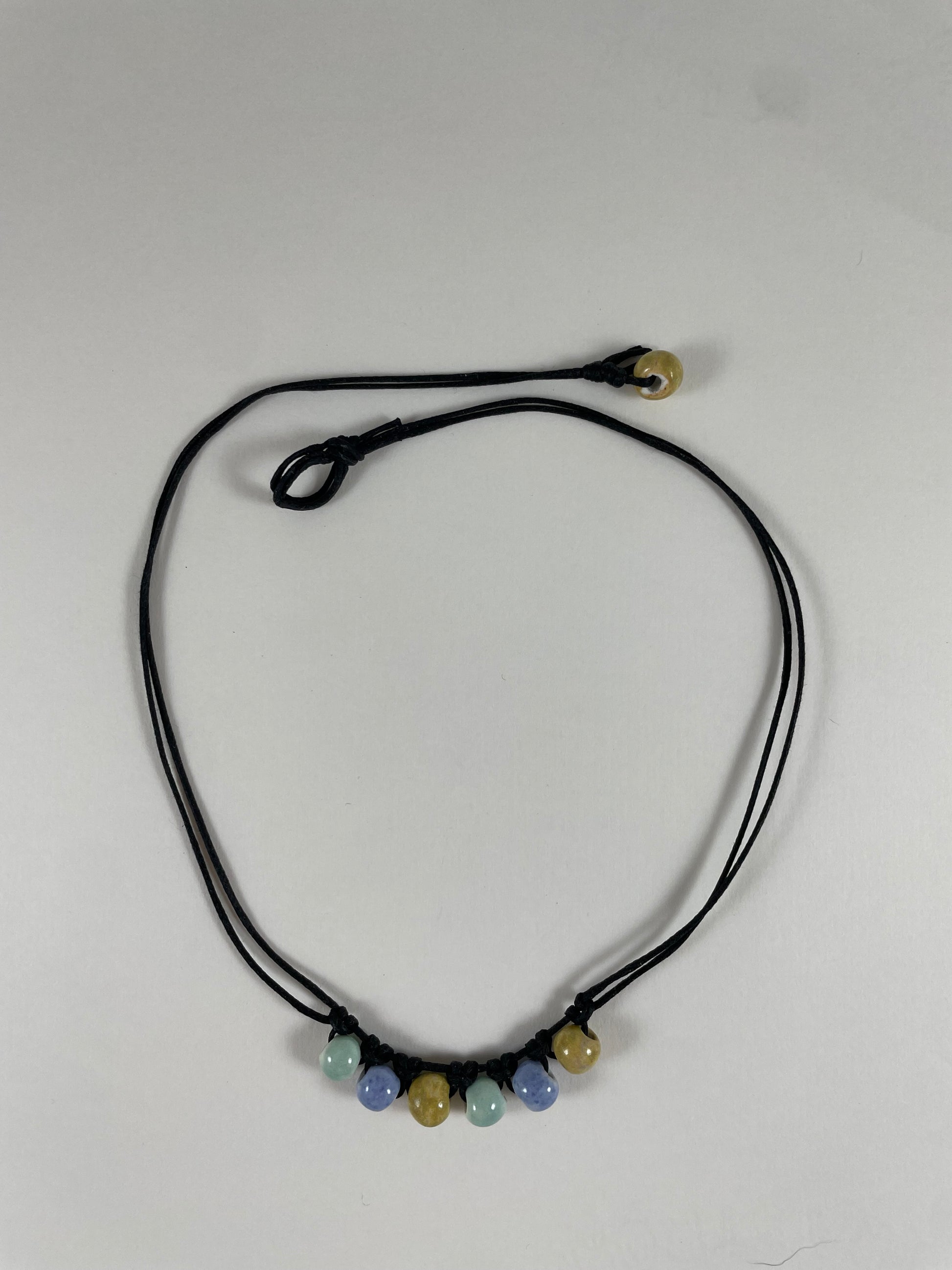 Short necklace laid flat on white background. Two strands of fine black string with six porcelain beads attached using simple macrame knots. Two beads are light blue, two are gold and two are light mint green. They are arranged in an alternating order. One end of necklace has a gold bead attached and the other end has a small loop for the bead to attach to.
