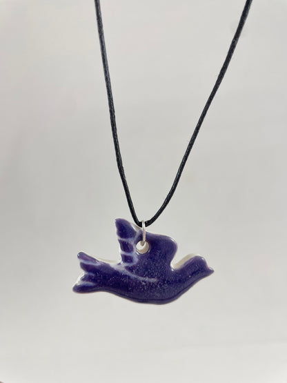 Necklace hanging against white background. Fine black thread with dove shaped pendant in dark lavender purple with fine white etching on wing and tail. Pendant is attached with small sterling silver jump ring.