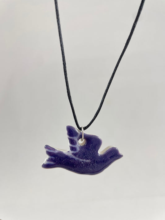 Necklace hanging against white background. Fine black thread with dove shaped pendant in dark lavender purple with fine white etching on wing and tail. Pendant is attached with small sterling silver jump ring.