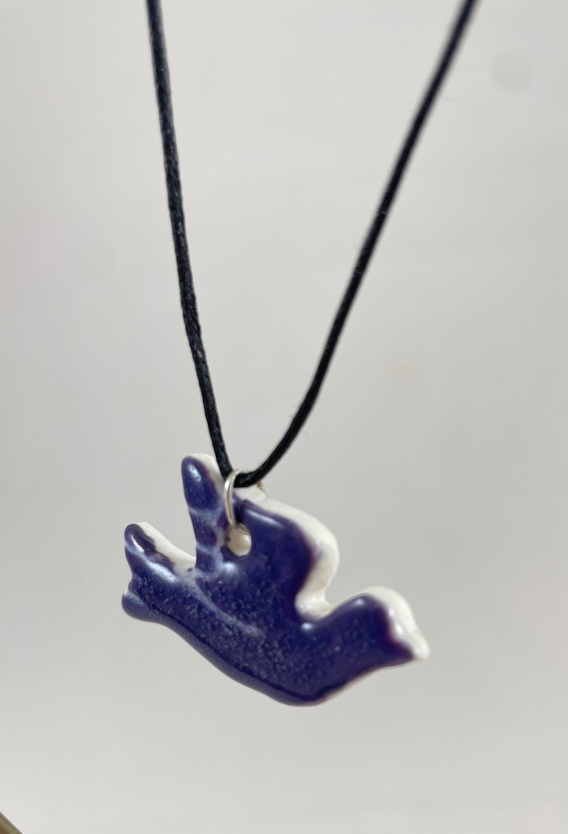 Necklace hanging against white background. Fine black thread with dove shaped pendant in deep lavender. Thin white etchings on wing and tail. Pendant is attached with small sterling silver jump ring. Side view. Pendant is made of thin porcelain.