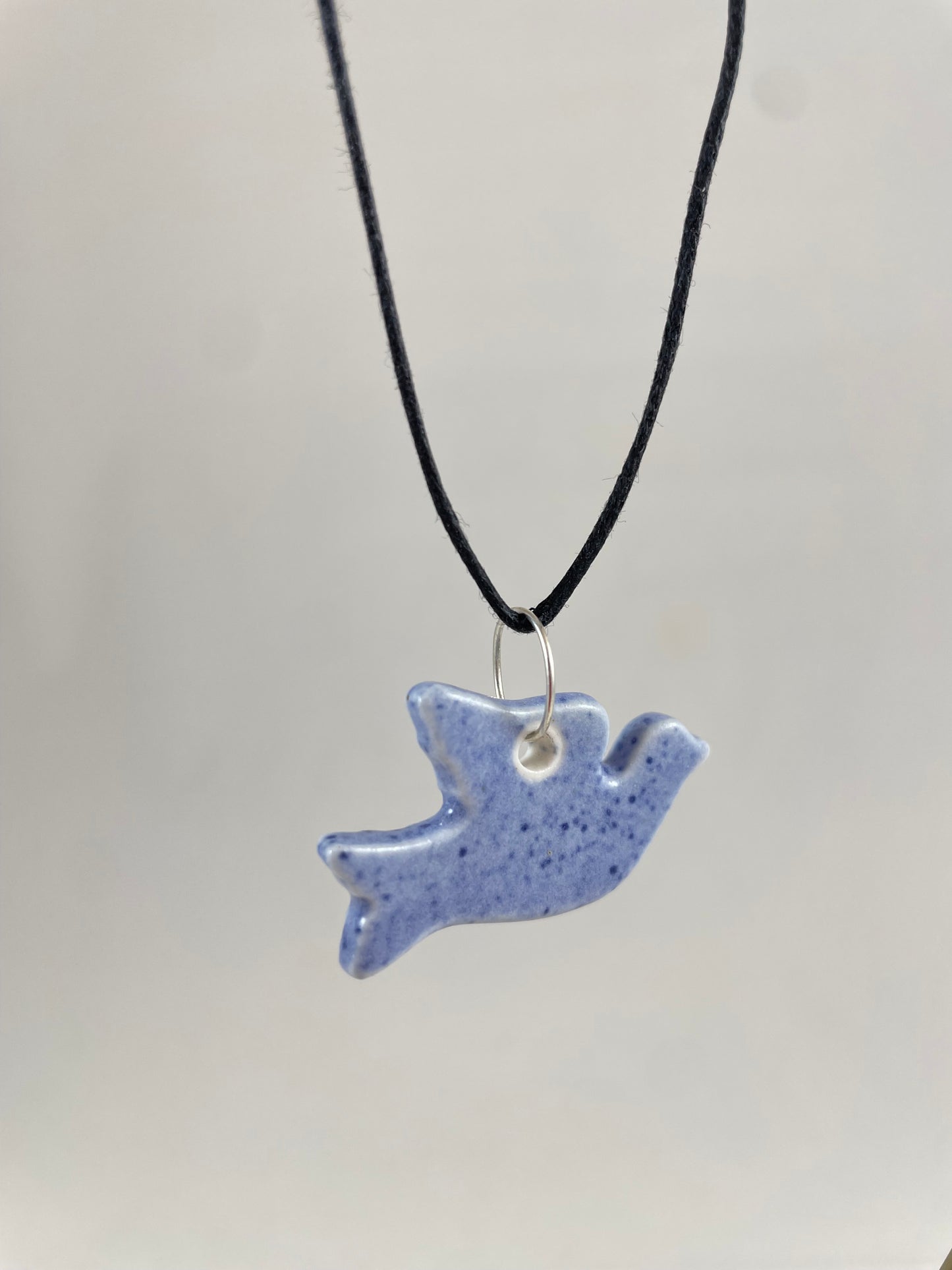 Necklace hanging against white background. Fine black thread with dove shaped pendant in pale blue with dark blue flecks. Pendant is attached with small sterling silver jump ring. Side view. Pendant is made of thin porcelain.