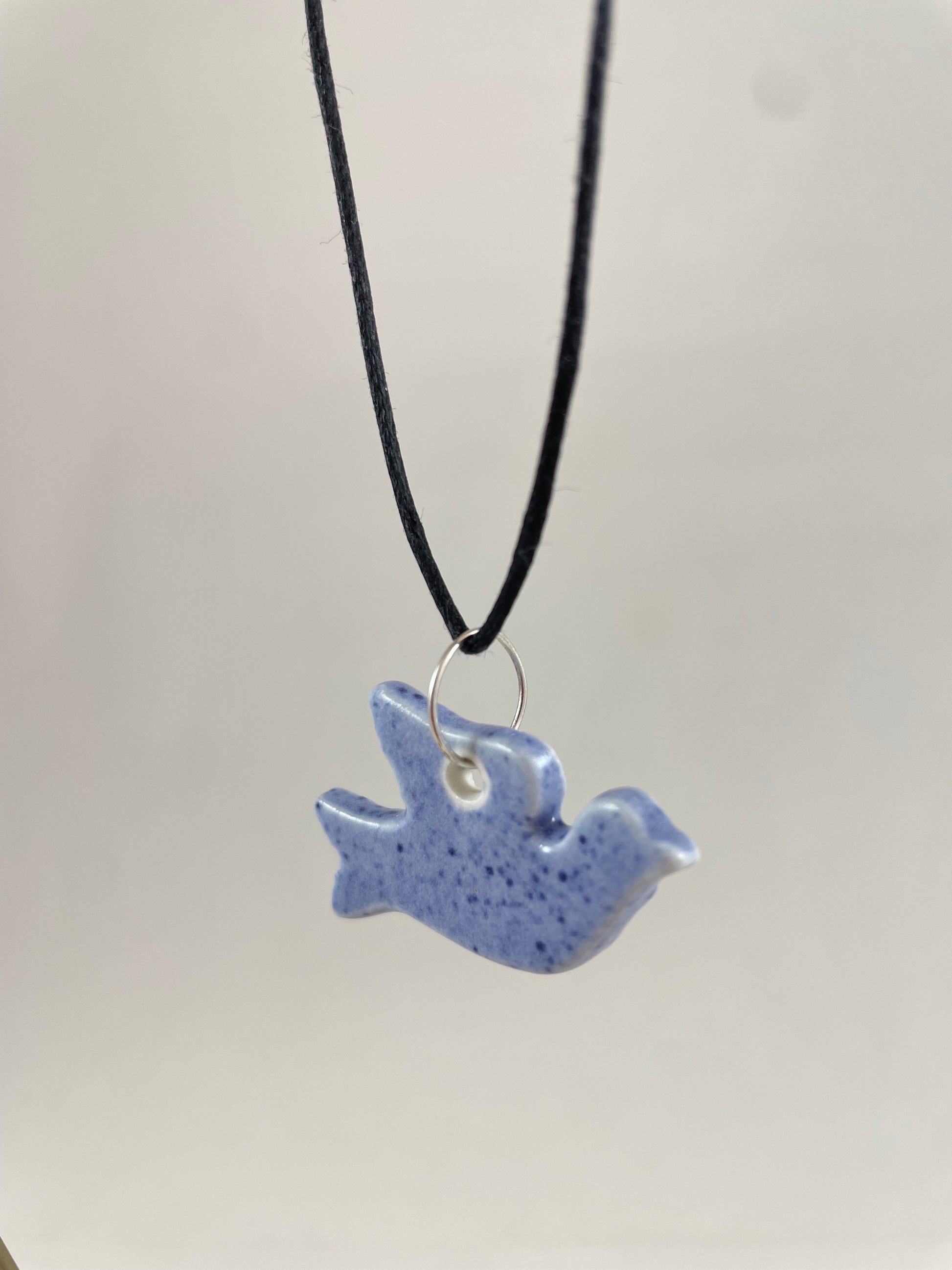 Necklace hanging against white background. Fine black thread with dove shaped pendant in pale blue with dark blue flecks. Pendant is attached with small sterling silver jump ring. Side view. Pendant is made of thin porcelain.