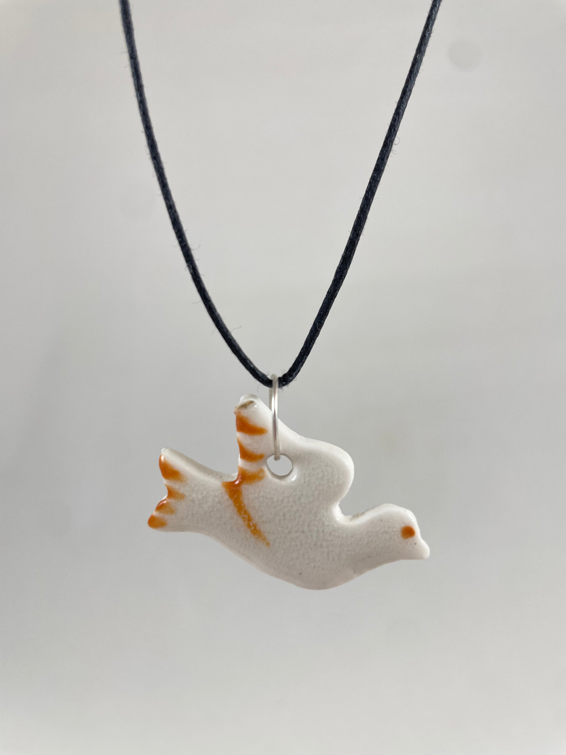 Necklace hanging against white background. Fine black thread with dove shaped pendant in white. Fine bright orange brush strokes on wing and tail. Pendant is attached with small sterling silver jump ring.