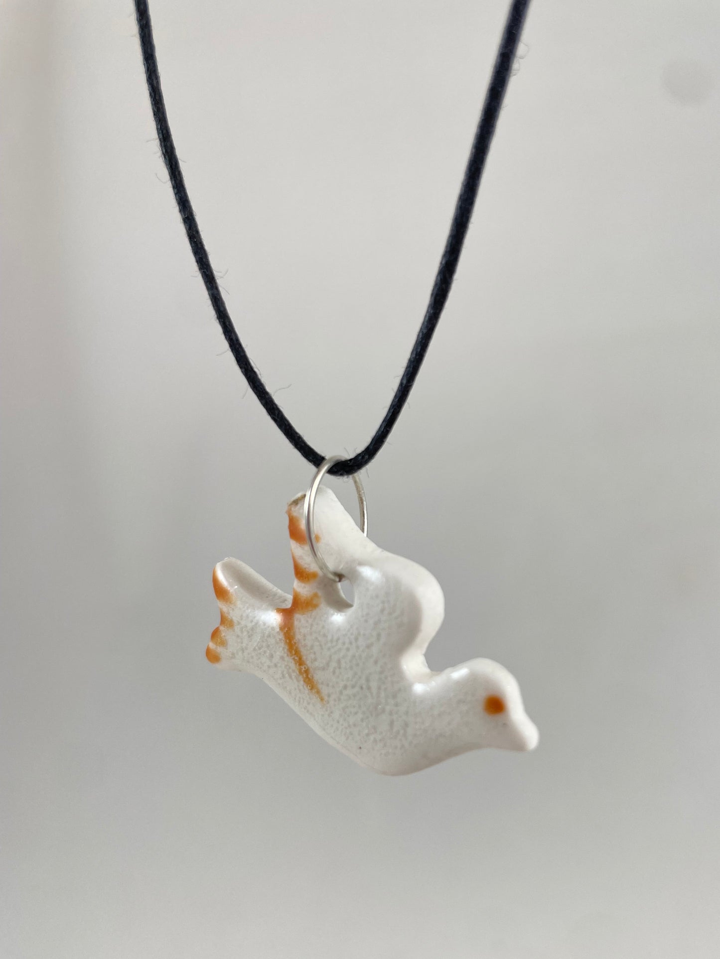 Necklace hanging against white background. Fine black thread with dove shaped pendant in white. Thin bright orange brush strokes on wing and tail. Pendant is attached with small sterling silver jump ring. Side view. Pendant is made of thin porcelain.
