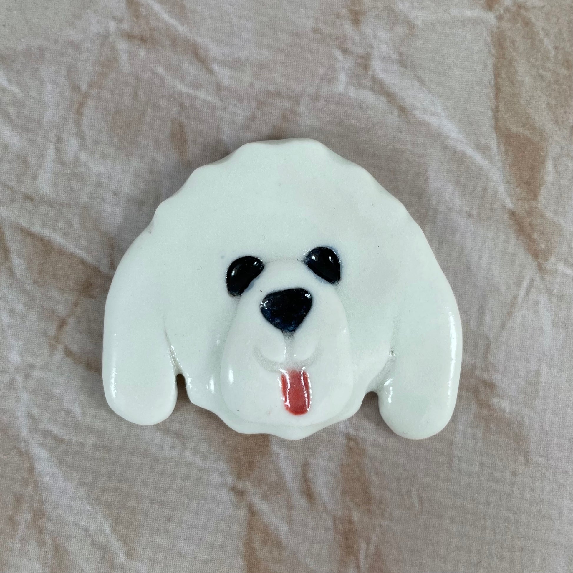 Dog's head in glossy white porcelain with long ears and pink tongue sticking out. Shiny black eyes and nose. Muzzle area is slightly raised to stand out. Photographed on paper that is printed with a crumpled effect.