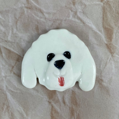 Dog's head in glossy white porcelain with long ears and pink tongue sticking out. Shiny black eyes and nose. Muzzle area is slightly raised to stand out. Photographed on paper that is printed with a crumpled effect.
