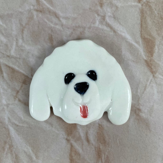 Dog's head in glossy white porcelain with long ears and pink tongue sticking out. Shiny black eyes and nose. Muzzle area is slightly raised to stand out. Photographed on paper that is printed with a crumpled effect.