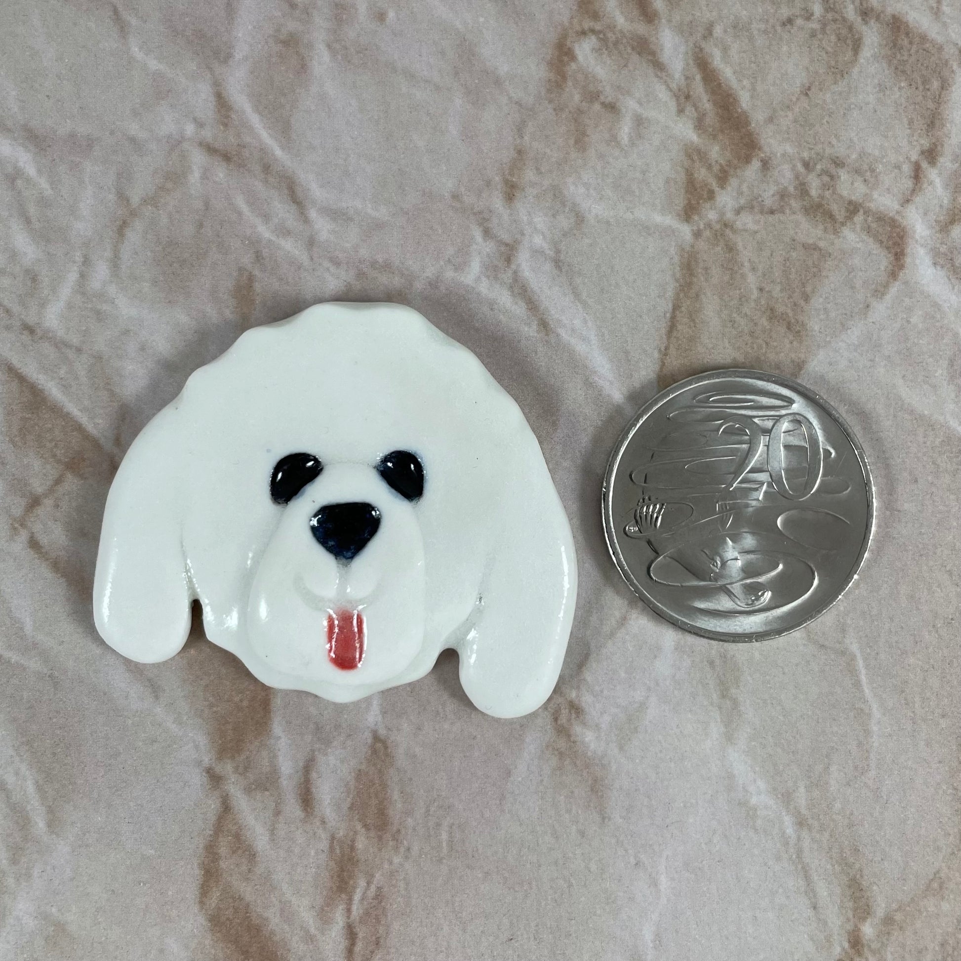 Dog's head in glossy white porcelain with long  ears and pink tongue sticking out. Shiny black eyes and nose. Muzzle area is slightly raised to stand out. Photographed next to 20 cent coin, which is smaller than brooch.