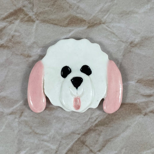Dog's head in glossy white porcelain with long pale pink ears and pink tongue sticking out. Shiny black eyes and nose. Muzzle area is slightly raised to stand out. Photographed on paper that is printed with a crumpled effect.