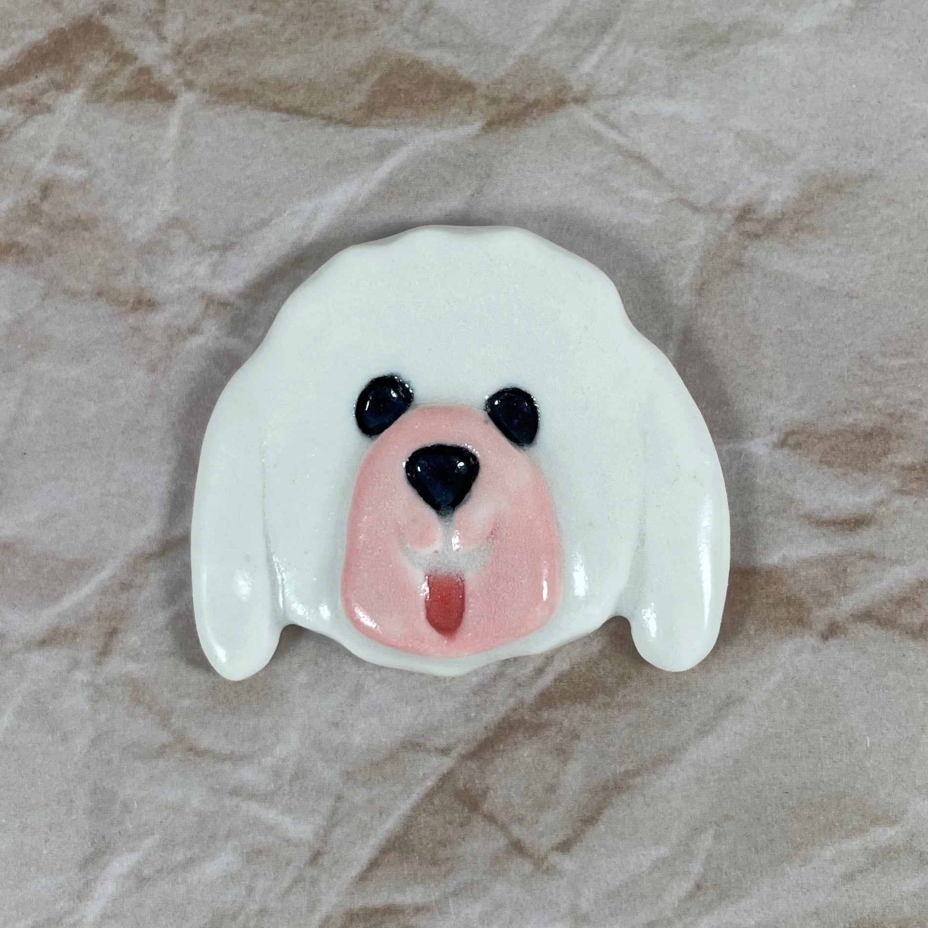 Dog's head in glossy white porcelain with long ears, pink tongue sticking out. Shiny black eyes and nose. Muzzle area is pale pink and is slightly raised to stand out. Photographed on paper that is printed with a crumpled effect.
