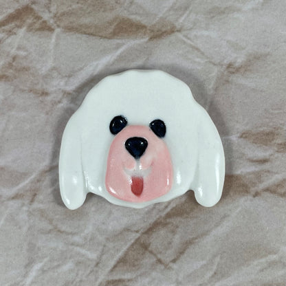 Dog's head in glossy white porcelain with long ears, pink tongue sticking out. Shiny black eyes and nose. Muzzle area is pale pink and is slightly raised to stand out. Photographed on paper that is printed with a crumpled effect.