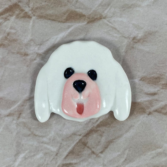 Dog's head in glossy white porcelain with long ears, pink tongue sticking out. Shiny black eyes and nose. Muzzle area is pale pink and is slightly raised to stand out. Photographed on paper that is printed with a crumpled effect.